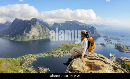L'escursionista gode di splendide vedute panoramiche di Reine e delle maestose isole Lofoten dalla cima di Reinebringen. I cieli limpidi e i paesaggi vivaci rendono questo luogo una fuga perfetta nella natura. Foto Stock
