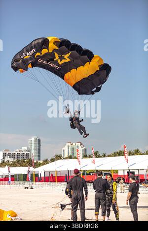 Membro della U.S. Army Parachute Team (USAPT), il sergente di prima classe Justin Brenner, vola con il suo paracadute su Miami Beach il 23 maggio 2025 prima dell'Hyundai Air and Sea Show a Miami, Florida. Miss Florida 2024, Casana Fink, si erge in primo piano con i membri della USAPT Tandem dopo aver completato un salto in tandem. I Cavalieri d'Oro conducono salti dimostrativi in tutto il paese per collegare il popolo americano all'esercito americano. (Foto dell'esercito degli Stati Uniti di SPC. Mari Kossman) Foto Stock