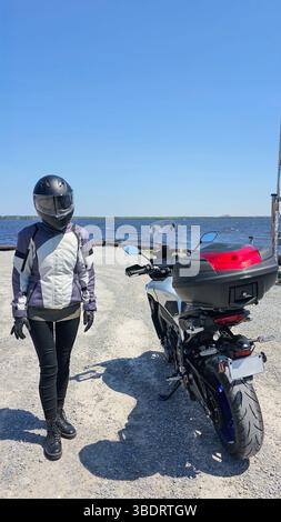 Una motociclista a tutto tondo si trova accanto alla sua moto su una strada panoramica lungo il lago. La giornata di sole e lo splendido paesaggio evocano un senso di libertà Foto Stock