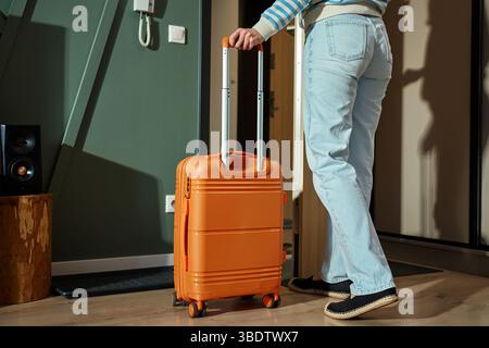 Persona che esce di casa con la valigia arancione. Viaggiatore femminile inizia il viaggio con i bagagli. Donna con un abbigliamento casual si trova vicino alla porta con sé la maniglia della valigia. TR Foto Stock