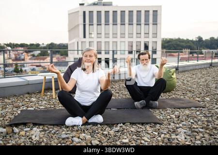 Coppie anziane che praticano meditazione yoga sul tetto in posa di loto all'aperto Foto Stock