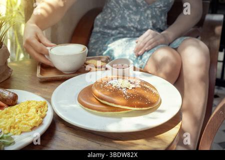 Una donna gode di una deliziosa colazione in un intimo caffè, caratterizzato da soffici pancake ricoperti di zucchero a velo e mandorle affettate. Tiene una tazza calda di Foto Stock