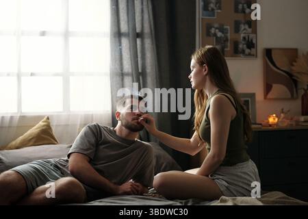 Coppia seduto sul letto in un'accogliente camera da letto dove godersi momenti di relax insieme. Camera decorata con foto e luce calda che aggiunge un'atmosfera intima Foto Stock