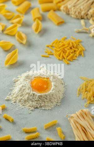 Diversi tipi di pasta che formano un cerchio intorno alla farina con un tuorlo d'uovo su sfondo grigio, pronto per essere cotto Foto Stock