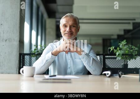 Felice e sorridente uomo d'affari professionista di mezza età, direttore esecutivo dell'azienda con indosso un abito blu seduto alla scrivania in ufficio e che lavora su un computer portatile Foto Stock