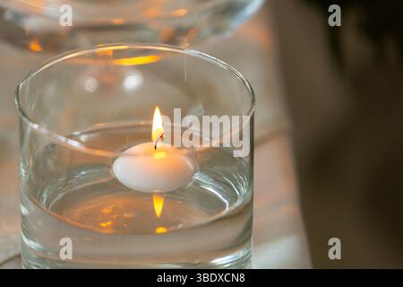 Primo piano di una piccola candela galleggiante bianca che brucia in un bicchiere d'acqua, creando un'atmosfera calda e invitante Foto Stock