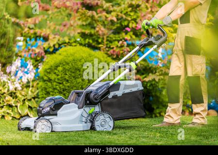Un giardiniere gestisce un rasaerba elettrico su erba ben tenuta circondata da aiuole colorate e vegetazione lussureggiante. Foto Stock
