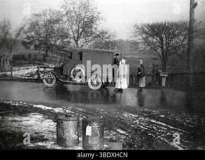 Si tratta di una foto originale e reale degli anni '1910 o '1920 che mostra una piovosa scena rurale lungo la strada. Due donne con cappotti lunghi e velo si trovano vicino a un camion per la consegna di pannelli all'inizio del XX secolo, probabilmente una Ford Model T o simili. La strada è bagnata e fangosa, con macchie di neve visibili, e una serie di lattine di latte d'epoca si trovano in primo piano, suggerendo il trasporto di latticini. Alberi senza fronzoli e una recinzione in legno aggiungono un'atmosfera rustica e ventosa. L'immagine cattura la vita quotidiana, i trasporti e le infrastrutture rurali nell'America dei primi anni del XX secolo. Foto Stock