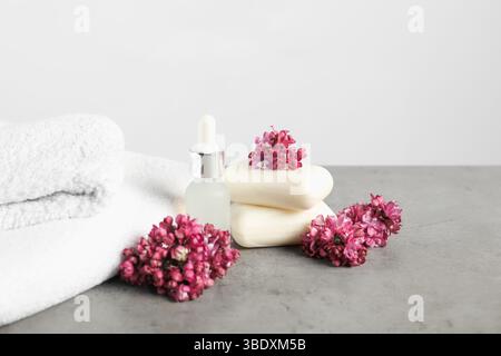 Pila di saponette, olio essenziale, asciugamani e fiori di lilla su un tavolo testurizzato grigio su sfondo chiaro, primo piano Foto Stock
