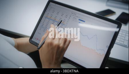 Trader di borsa femminile monitoraggio e analisi delle azioni in tempo reale, grafici di mercato nell'app di trading online con tablet e stilo digitali. Agente di investimento professionale che lavora in ufficio. Primo piano. Foto Stock