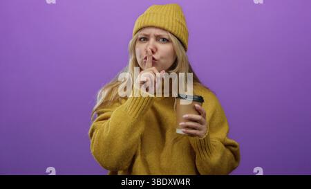 Una donna in giallo sembra curiosa con un dito sulle labbra che tiene il caffè contro una vivace parete viola. Foto Stock