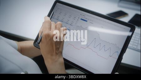 Trader di borsa femminile monitoraggio e analisi delle azioni in tempo reale, grafici di mercato nell'app di trading online con tablet e stilo digitali. Agente di investimento professionale che lavora in ufficio. Primo piano. Foto Stock
