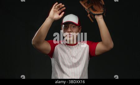 Il giovane cinese in uniforme da baseball posa con sicurezza con il guanto su sfondo nero isolato. Foto Stock