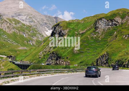 Le auto guidano la Grossglockner High Alpine Road montagne colline verdi cime blu cielo sfondo. Il percorso tortuoso attraverso il paesaggio montano offre una vista panoramica Foto Stock