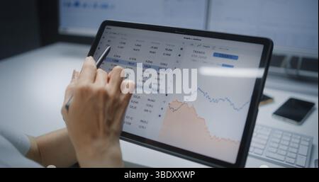 Analista finanziaria donna che controlla le azioni in tempo reale, grafici del mercato Exchange nell'app mobile di trading online su tablet computer con penna digitale. Donna d'affari che lavora nell'ufficio dell'agenzia finanziaria. Primo piano Foto Stock