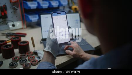 Uomo che controlla azioni in tempo reale, grafici del mercato Exchange su smartphone e laptop. Dipendente che effettua investimenti in criptovaluta durante l'orario di lavoro in officina, combinando lavoro e Trading online. Primo piano Foto Stock
