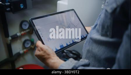 Dipendente che controlla azioni in tempo reale e grafici Exchange Market utilizzando un computer tablet digitale. Uomo che effettua investimenti durante l'orario di lavoro in officina, combinando lavoro e Trading online. Primo piano. Foto Stock