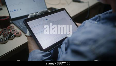 Uomo che controlla azioni in tempo reale e grafici Exchange Market utilizzando tablet e laptop digitali. Dipendenti che effettuano investimenti durante l'orario di lavoro in officina, combinando il lavoro con il trading online. Primo piano. Foto Stock