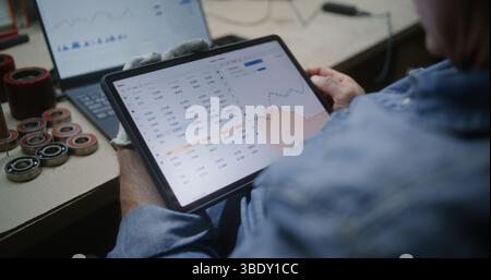 Uomo che controlla azioni in tempo reale e grafici Exchange Market utilizzando tablet e laptop digitali. Dipendenti che effettuano investimenti durante l'orario di lavoro in officina, combinando il lavoro con il trading online. Primo piano. Foto Stock
