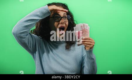 Una donna di mezza età che tiene in mano banconote in corona islandese guarda in lontananza contro una vivace parete verde, trasmettendo curiosità ed eccitazione. Foto Stock