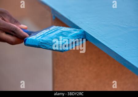pennellate con vernice blu tenuta da pittore africano, dipingendo una scrivania in legno in una falegnameria Foto Stock