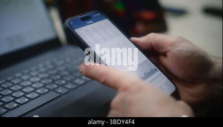 Uomo che controlla le azioni in tempo reale e i grafici del mercato di Exchange su smartphone e laptop, guarda la lezione di trading online su tablet. Dipendenti che effettuano investimenti durante l'orario di lavoro in officina. Primo piano. Foto Stock