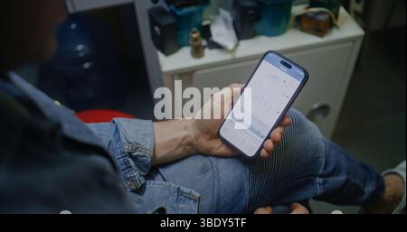 Uomo che controlla le azioni in tempo reale e i grafici del mercato Exchange tramite smartphone. Dipendente che effettua investimenti in criptovaluta durante l'orario di lavoro in officina, combinando lavoro e Trading online. Primo piano Foto Stock