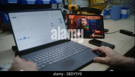 Uomo che controlla azioni in tempo reale e grafici del mercato Exchange utilizzando un laptop e guarda il tutorial sul trading online su un tablet digitale. Attività di trading di apprendimento remoto dei dipendenti durante l'orario di lavoro in officina. Primo piano. Foto Stock