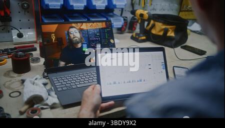 Dipendente che guarda la lezione di trading online sul laptop, controlla le azioni in tempo reale, Scambio grafici del mercato con smartphone e tablet digitali. Uomo Remote Learning Trading che lavora in officina. Primo piano. Foto Stock