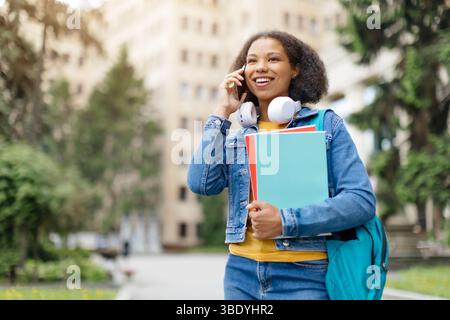 Una studentessa afroamericana sorridente parla al cellulare mentre cammina all'aperto Foto Stock