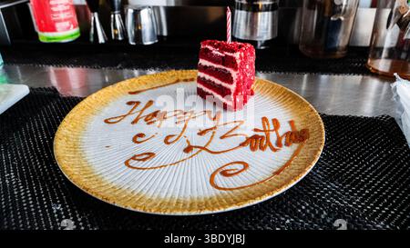 Un pezzo di torta di velluto rosso con una candela accesa siede su un piatto dove viene scritto "Happy Birthday". Una superficie scura e ruvida funge da sfondo Foto Stock