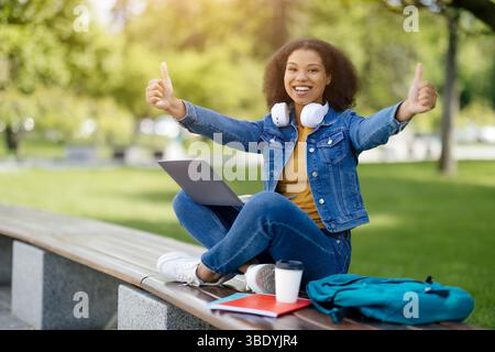 Studentessa di colore che posa con un notebook e gesti su pollice all'aperto Foto Stock