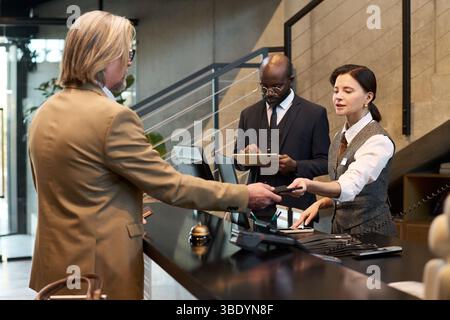 Uomo d'affari in costume beige che interagisce con il personale della reception mentre effettua il check-in alla reception. L'addetto alla reception scansiona la carta mentre un altro membro del personale prende appunti sugli appunti Foto Stock