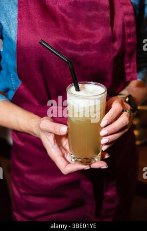 Un barista in un grembiule Bordeaux che tiene un rinfrescante cocktail di agrumi ghiacciato in un alto bicchiere con paglia nera. Foto Stock