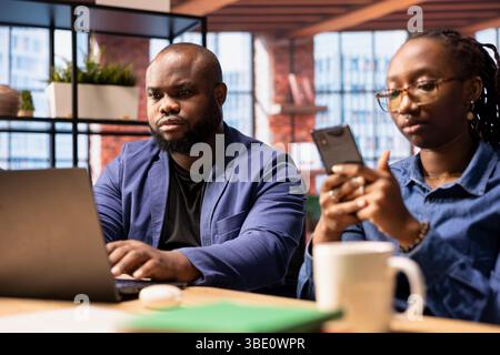 Una coppia afroamericana in un appartamento moderno collabora a un progetto di consulenza, utilizzando la tecnologia per migliorare la propria produttività. L'uomo lavora al portatile mentre il suo partner risponde alle e-mail al telefono. Foto Stock