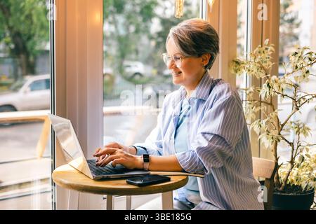 Donna anziana sorridente che lavora a distanza su un computer portatile al tavolo della finestra con telefono e vegetazione Foto Stock
