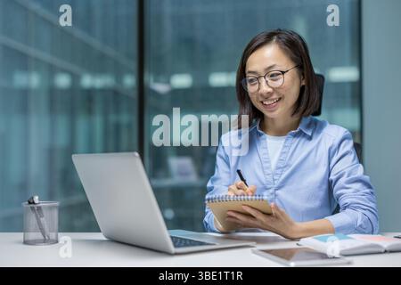 Una donna asiatica sorridente con gli occhiali lavora alla sua scrivania, prendendo appunti vicino a un portatile in un ambiente moderno di ufficio. Foto Stock