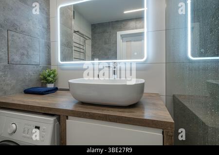 Bagno contemporaneo con lavandino bianco su ripiano in legno, doccia in vetro con gocce di pioggia e pareti piastrellate grigie. Foto Stock