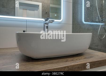 Bagno contemporaneo con lavandino bianco su ripiano in legno, doccia in vetro con gocce di pioggia e pareti piastrellate grigie. Foto Stock