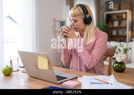 Giovane donna seduta alla scrivania di casa, indossa cuffie e abiti casual, concentrandosi su documenti e appunti, lavorando da remoto o studiando, circondati Foto Stock