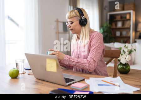Giovane donna seduta alla scrivania di casa, indossa cuffie e abiti casual, concentrandosi su documenti e appunti, lavorando da remoto o studiando, circondati Foto Stock