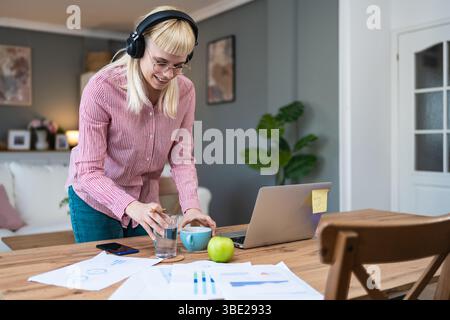 Giovane donna seduta alla scrivania di casa, indossa cuffie e abiti casual, concentrandosi su documenti e appunti, lavorando da remoto o studiando, circondati Foto Stock