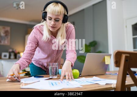 Giovane donna seduta alla scrivania di casa, indossa cuffie e abiti casual, concentrandosi su documenti e appunti, lavorando da remoto o studiando, circondati Foto Stock