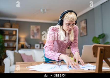 Giovane donna seduta alla scrivania di casa, indossa cuffie e abiti casual, concentrandosi su documenti e appunti, lavorando da remoto o studiando, circondati Foto Stock