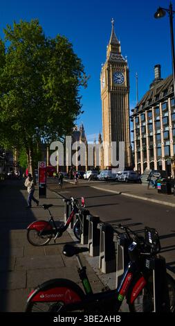 Big Ben dal Victoria Embankment con cielo blu, alberi frondosi e traffico leggero con biciclette elettriche parcheggiate in primo piano Foto Stock