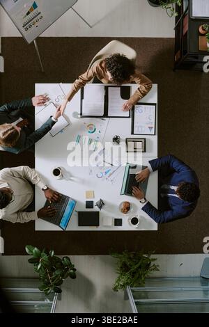 Quattro professionisti seduti al tavolo dell'ufficio a discutere di lavoro utilizzando computer portatili e documenti. La vista dall'alto verso il basso fornisce una prospettiva dell'area di lavoro collaborativa Foto Stock