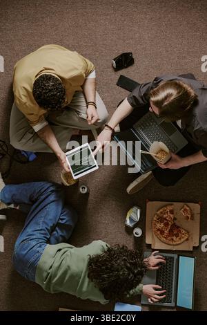 Gruppo di colleghi che discutono del progetto mentre mangiano cibo e utilizzano vari dispositivi elettronici nell'ambiente di lavoro con atmosfera rilassata e cas Foto Stock