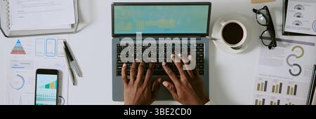 Vista dall'alto delle mani che lavorano sul laptop, analizzano documenti finanziari e grafici, con caffè, smartphone e occhiali disposti in modo ordinato sulla scrivania Foto Stock