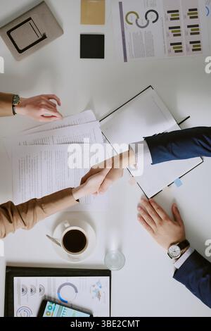 Gli uomini d'affari stringono la mano al tavolo delle riunioni con documenti, grafici e una tazza di caffè. Immagine dell'accordo, della collaborazione e del successo Foto Stock