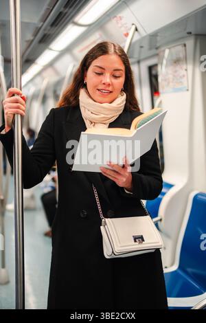 Una donna concentrata immersa nella lettura di un libro accattivante mentre si trova in una metropolitana, che mostra il suo piacere di leggere la letteratura durante i suoi spostamenti Foto Stock
